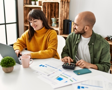 couple sitting at table looking at laptop, papers, and calculator
